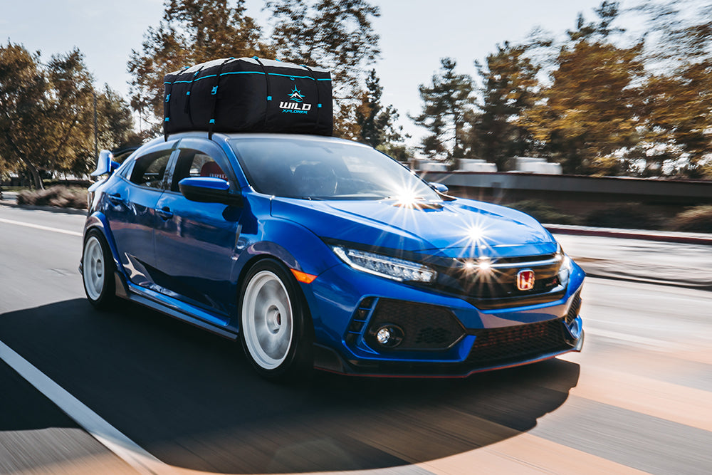Blue Honda car with a roof bag driving on a road with trees in the background
